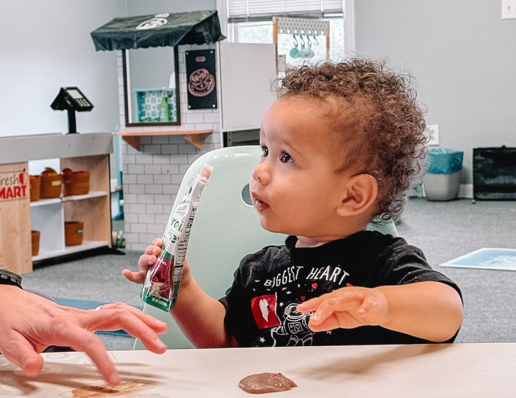 Young Black baby with curly hair holding a squeezy pouch, with some spilled on the table, looking up at a feeding therapist.