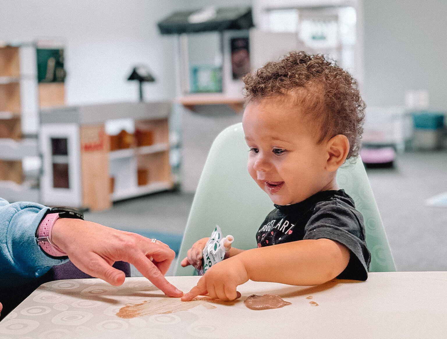 Young Black baby with curly hair holding a squeezy pouch, with some spilled on the table, looking up at a feeding therapist.